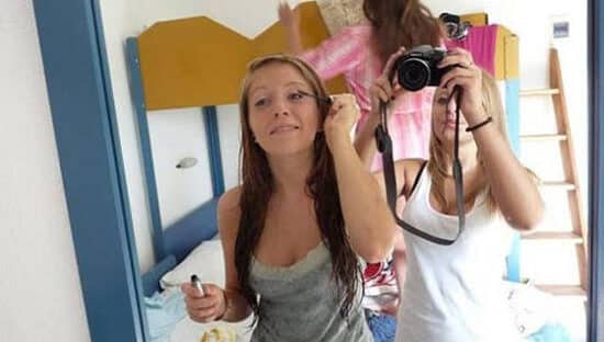Dwie dziewczyny robią selfie w pokoju, jedna trzyma telefon, druga uśmiecha się do aparatu.
