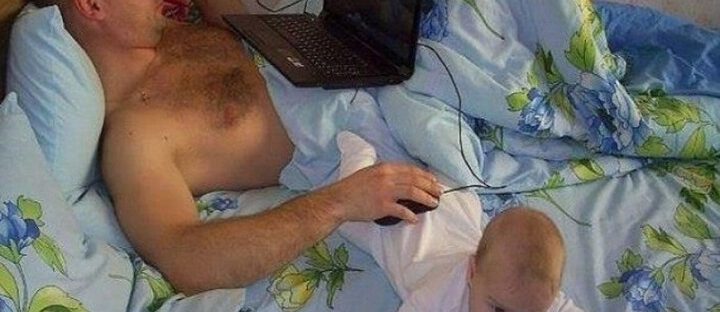 Ojciec leży na łóżku z laptopem, dziecko na podłodze obok.