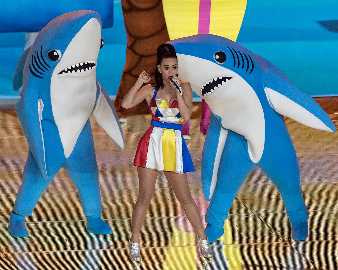 Katy Perry podczas Super Bowl 2015 w kolorowej sukience, pomiędzy dwoma dużymi niebieskimi rekinami