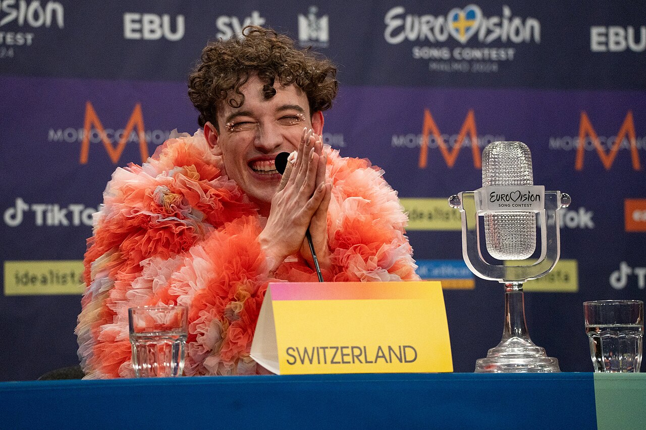 Nemo w różowo-pomarańczowym stroju na konferencji zwycięzcy Eurowizji 2024, obok stoi szklana statuetka mikrofonu