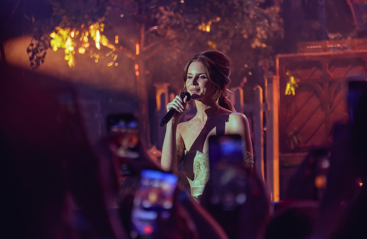 Lana Del Rey śpiewa solo na Wembley 2025 Lana Del Rey śpiewa do mikrofonu na scenie Wembley Stadium w ciepłym oświetleniu, na pierwszym planie telefony fanów