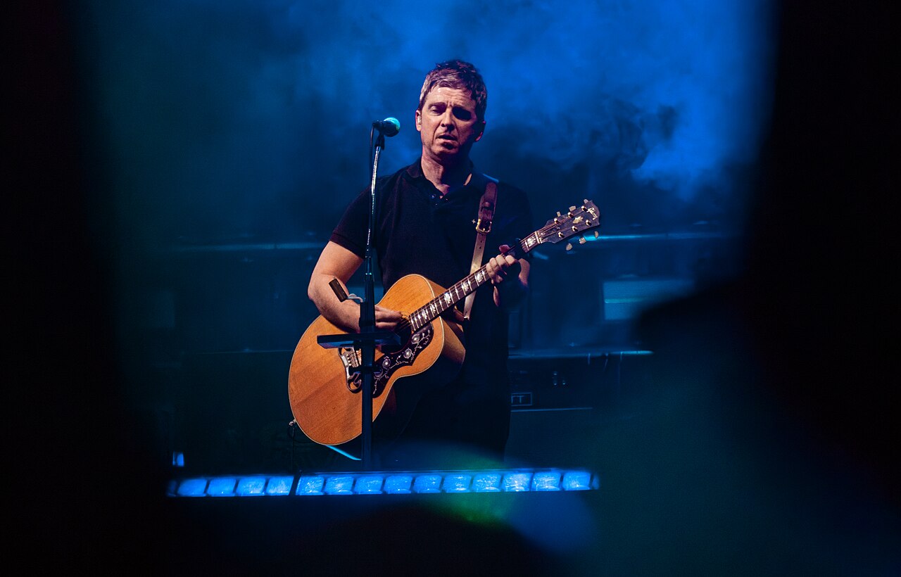 Noel Gallagher gra na gitarze akustycznej podczas Oasis Live 25 Noel Gallagher śpiewa i gra na akustycznej gitarze przed mikrofonem w niebieskim świetle koncertowym podczas Oasis Live 25 na Wembley