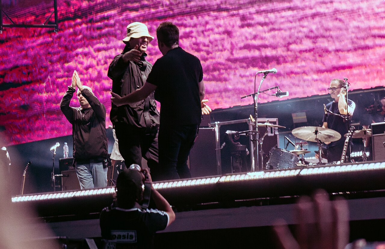 Bracia Liam i Noel Gallagher stoją naprzeciwko siebie podczas koncertu Oasis na Wembley Stadium 3 sierpnia 2025, w tle perkusista zespołu