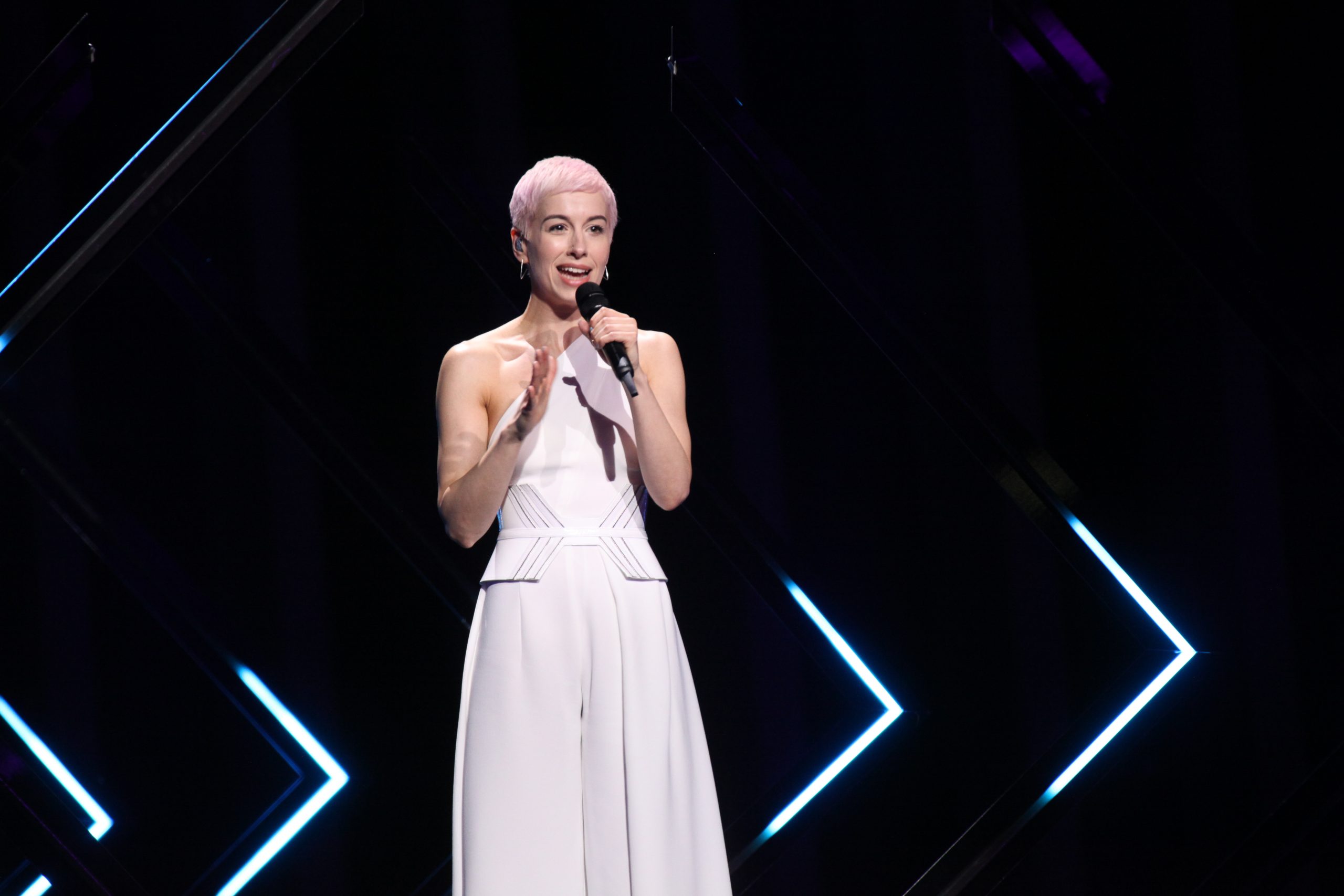 SuRie na próbie Eurowizji 2018 - zbliżenie SuRie z krótkimi blond włosami i mikrofonem na próbie Eurowizji 2018, w białym stroju i pośród neonowej scenografii