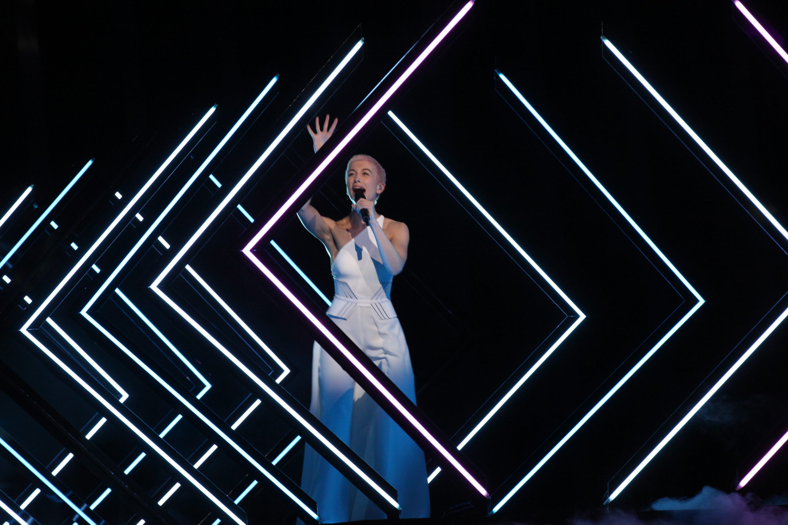 SuRie w białym kombinezonie śpiewa Storm w geometrycznej świetlnej scenografii podczas próby Eurowizji 2018